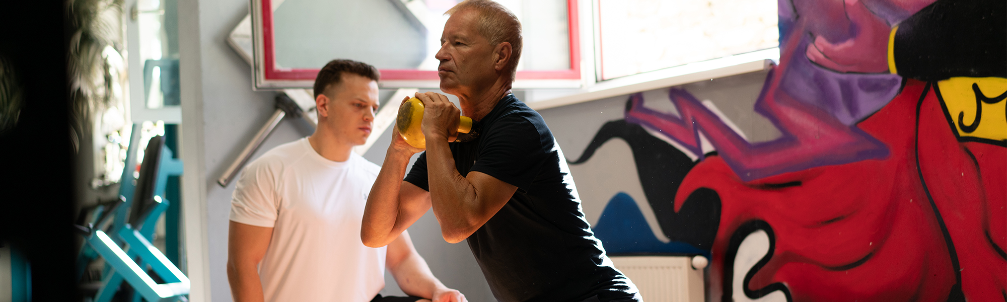 Personaltrainer in wei&szlig;en Shirt, achtet auf die Form eines &auml;lteren Herren bei der Kniebeuge mit einer gelben Kettlebell.