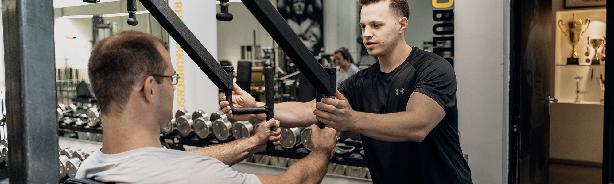 Ein Personaltrainer assistiert einem jungen Mann bei einer Brust&uuml;bung in einer Maschine.
