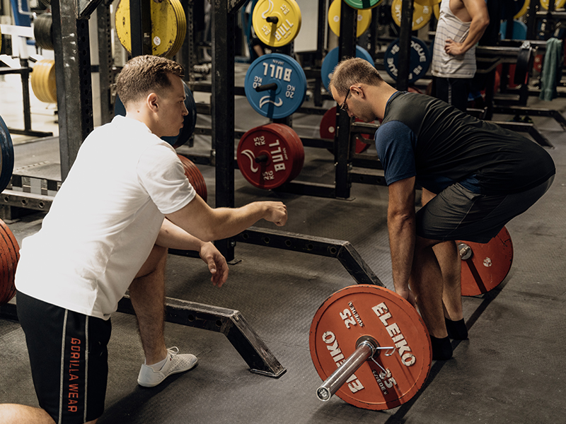 Personaltrainer in wei&szlig;em Shirt coacht einem jungen Mann in einem Gym in der Ausgangsposition der &Uuml;bung "Deadlifts".