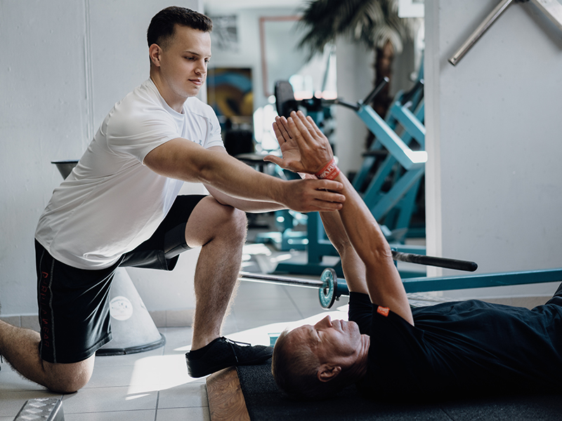 Personaltrainer unterst&uuml;tzt einen &auml;lteren, am Boden liegenden Herren bei der Armposition einer Mobilisations&uuml;bung.